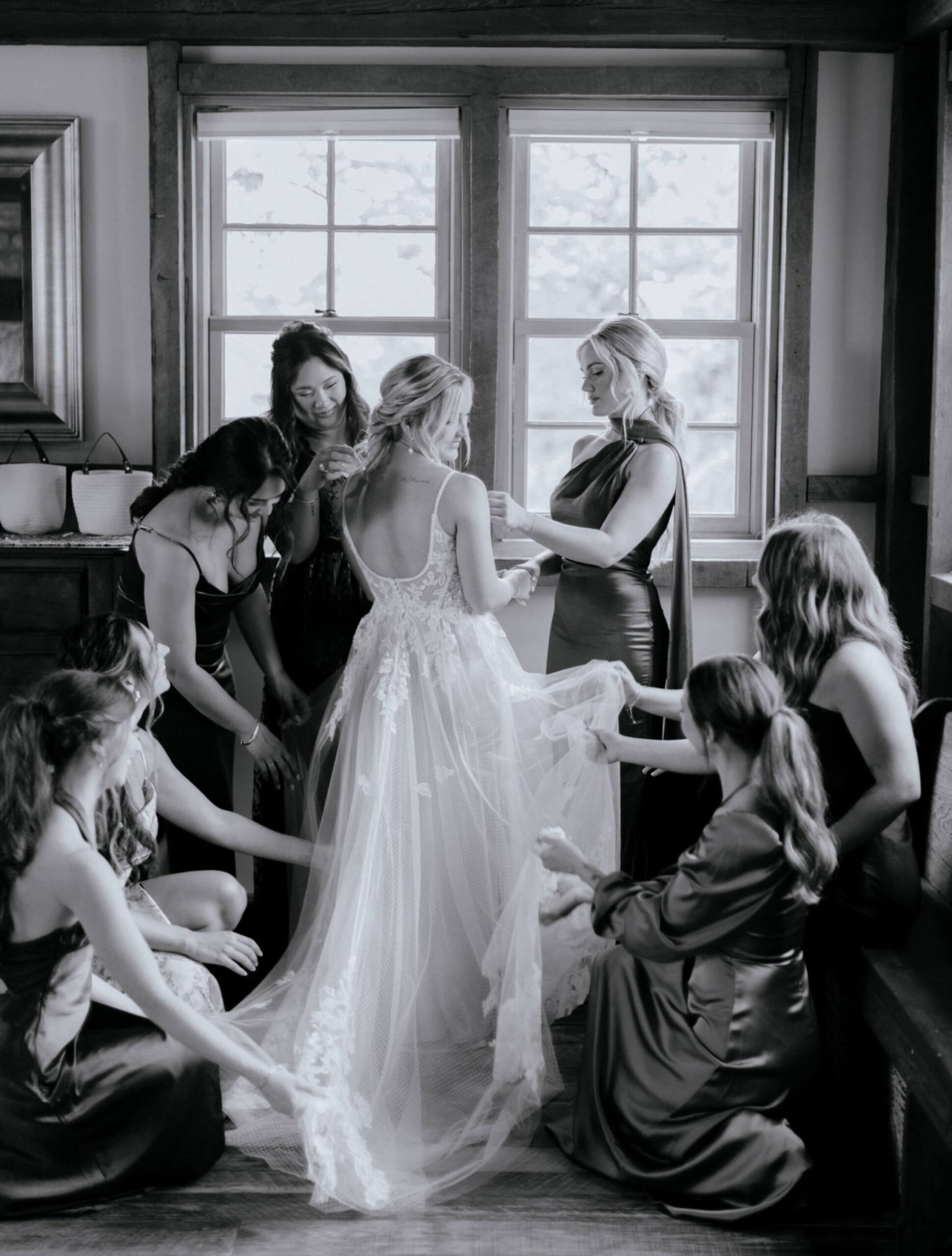 A bride stands in a room while bridesmaids in dark dresses help arrange her gown and veil. Natural light comes through the windows behind them. The image is in black and white.