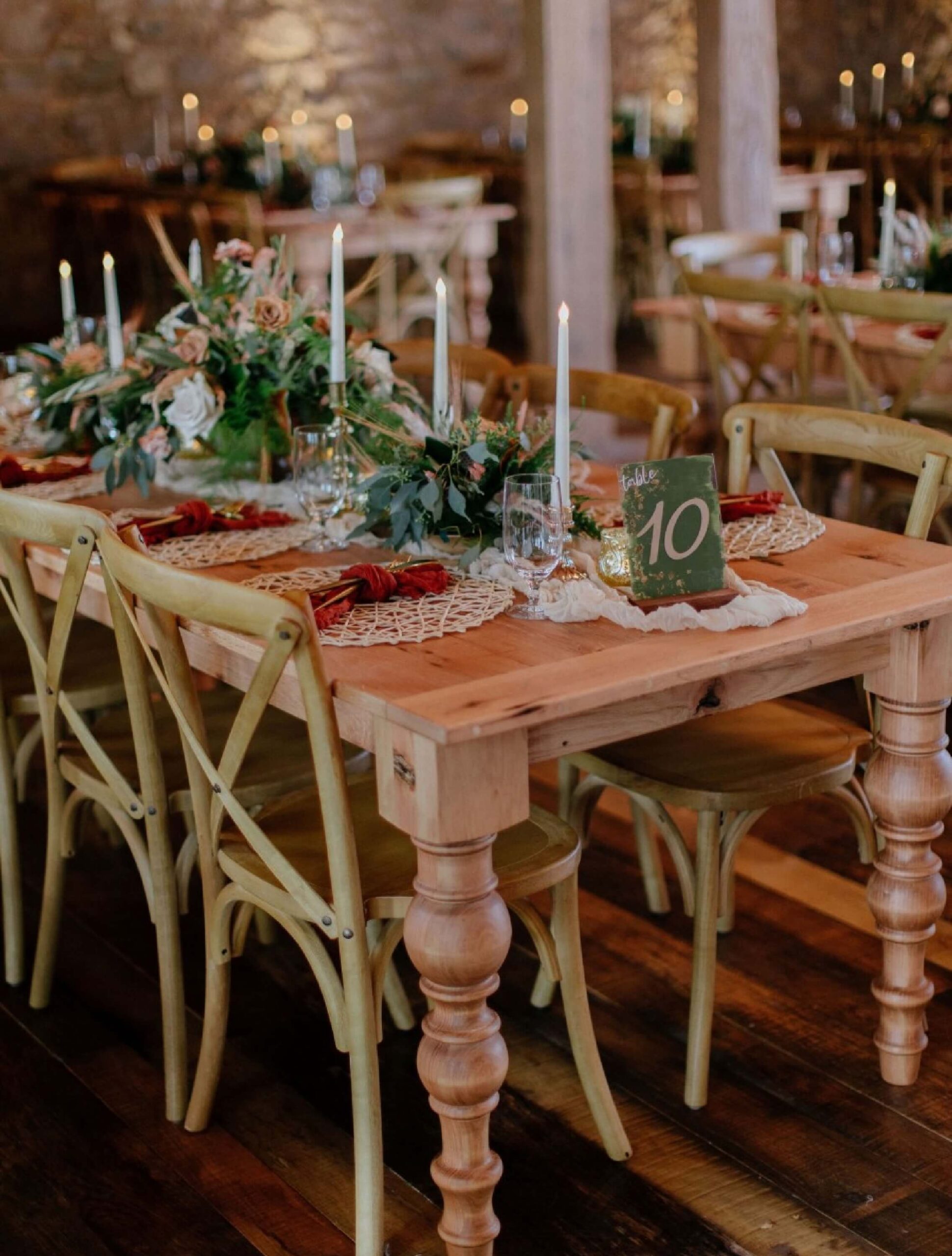 A rustic wooden table set for a meal with floral centerpieces, candles, place settings, and a table number 10 card, surrounded by wooden chairs in a warmly lit room.