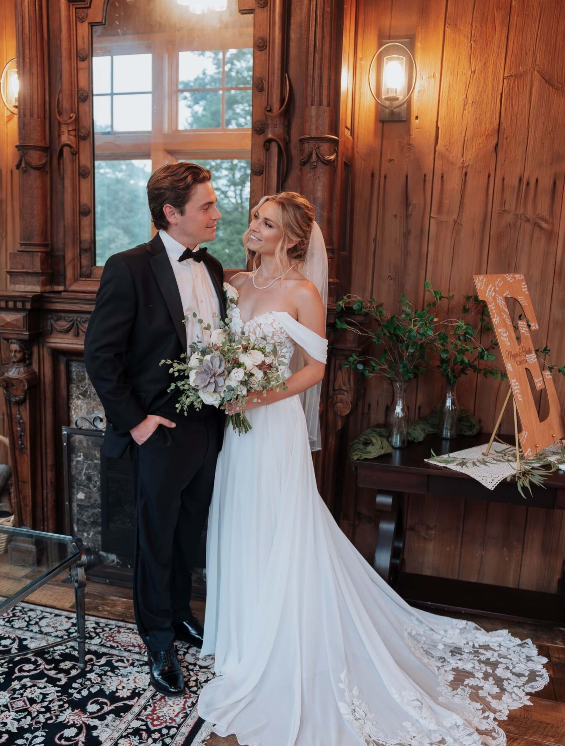 A bride in a white gown and groom in a black tuxedo stand together indoors, surrounded by wood paneling and decor, posing for a wedding photo.