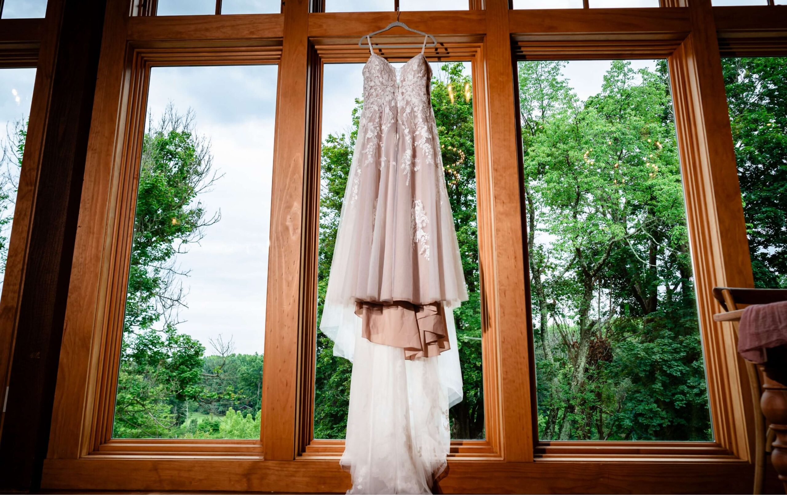 A white wedding dress with lace details hangs on a wooden window frame, with green trees and a cloudy sky visible outside.