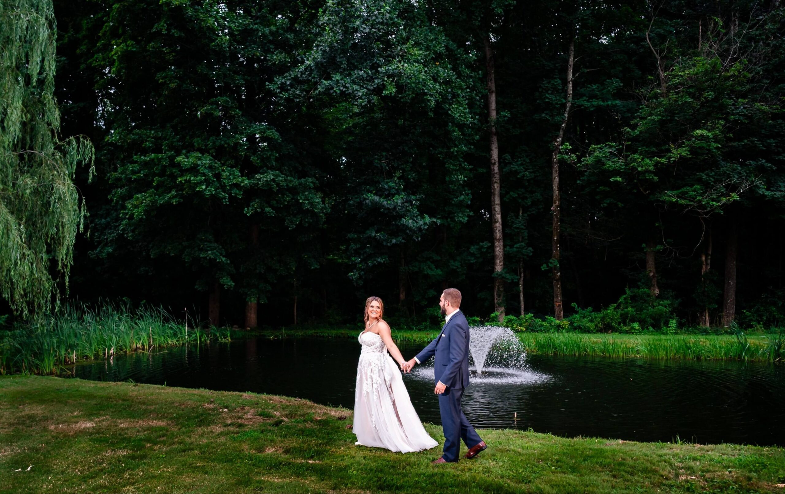 A bride and groom hold hands and walk on grass near a pond with a fountain, surrounded by dense trees.