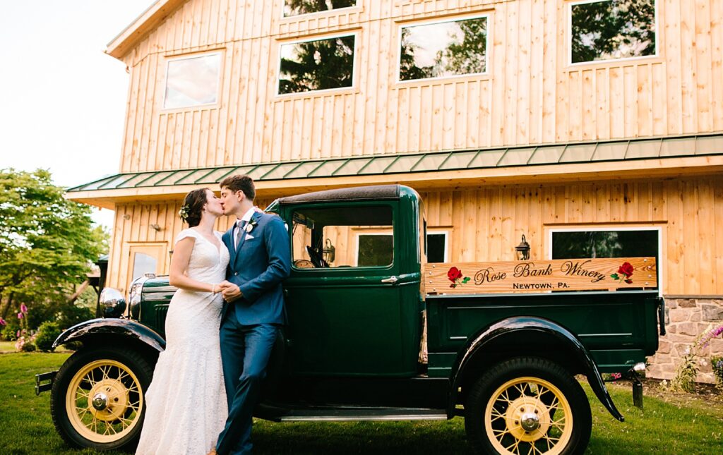 A bride and groom kiss beside a vintage green truck labeled “Rose Bank Winery” in front of a wooden building.