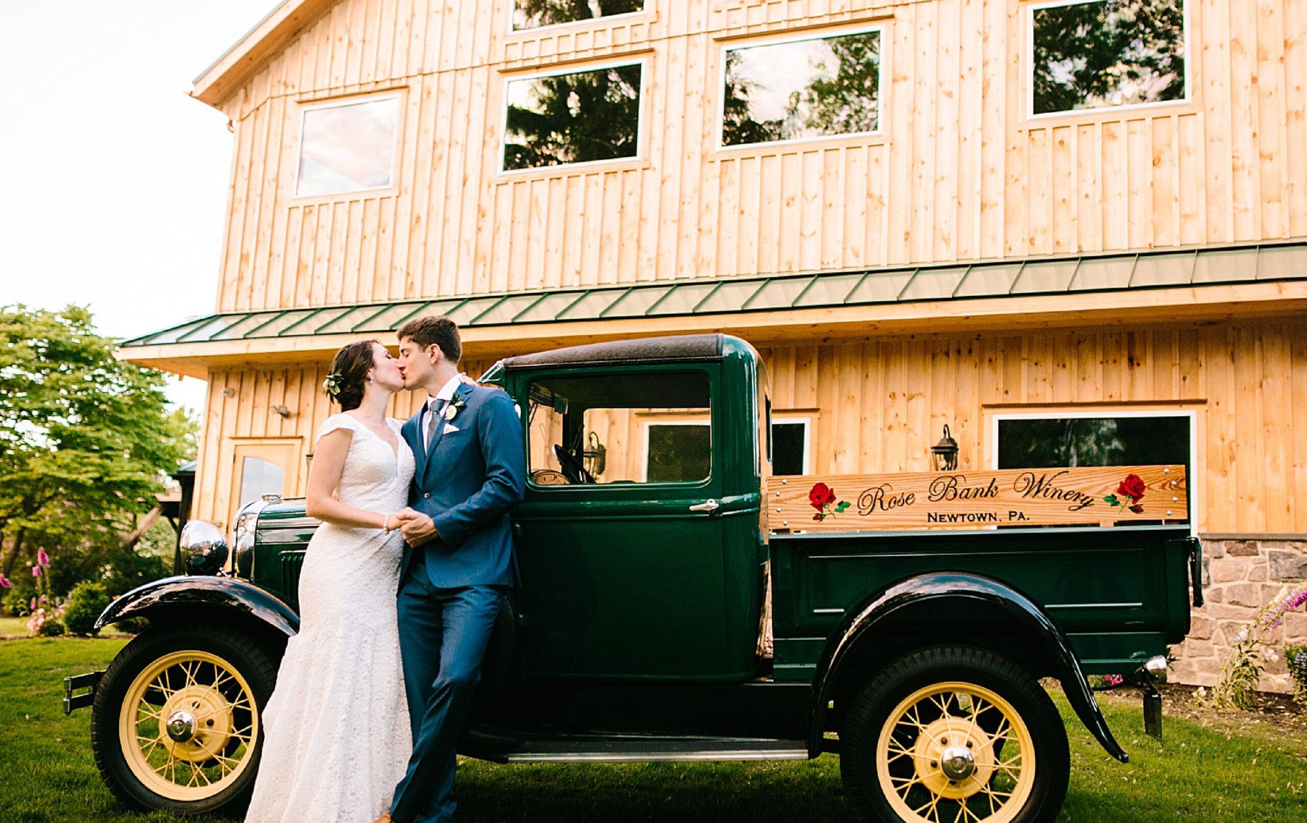 A bride and groom kiss beside a vintage green truck labeled “Rose Bank Winery” in front of a wooden building.