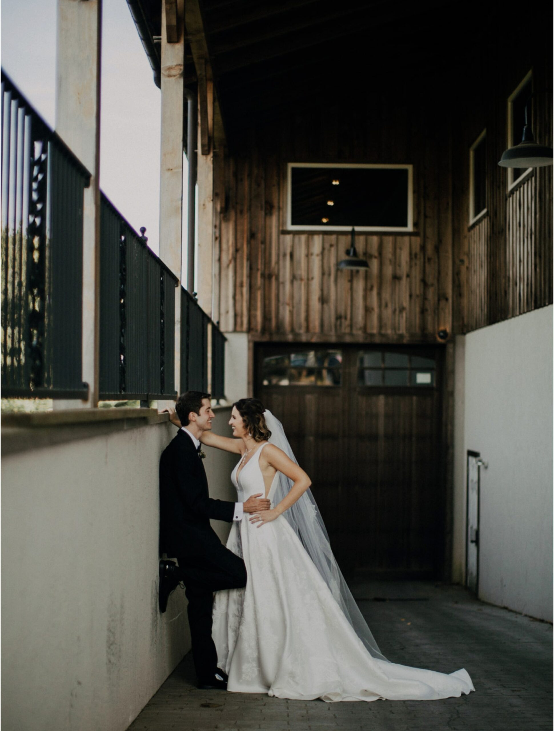 A bride and groom stand close together in an outdoor, covered walkway next to a wooden building, looking at each other.