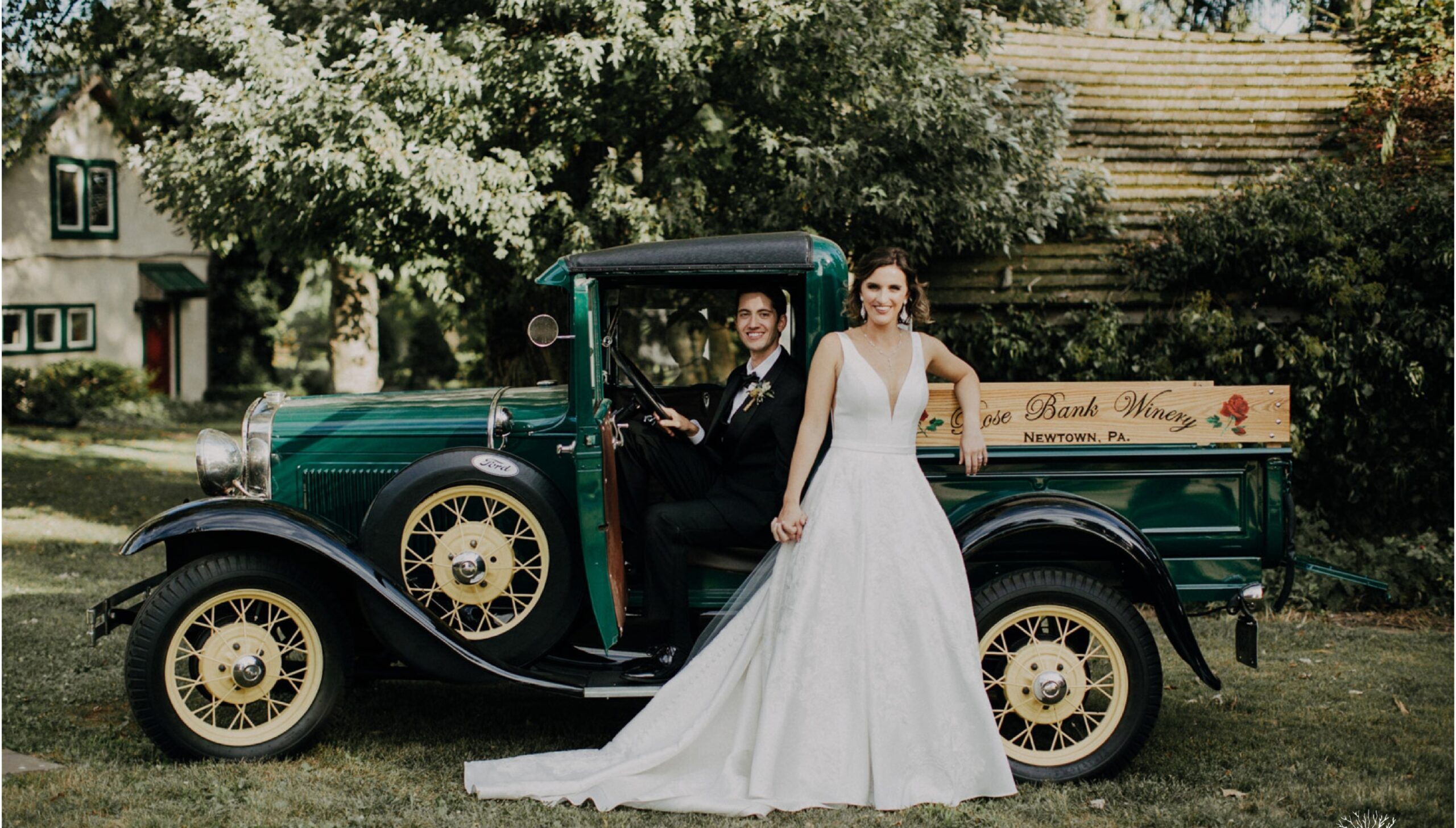 A bride and groom pose outdoors beside a vintage green truck; the groom sits in the driver’s seat while the bride stands holding his hand.