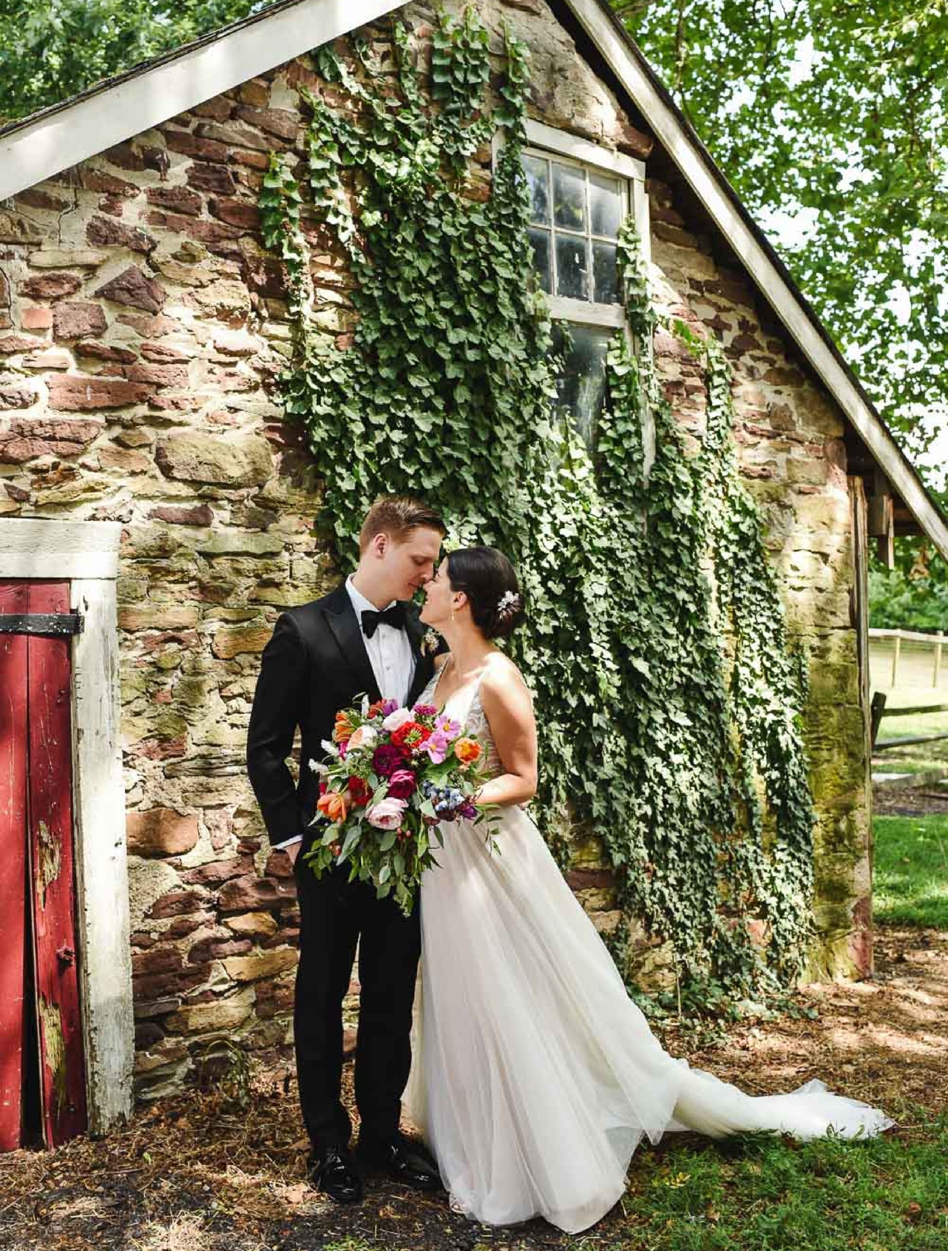 A bride and groom stand close together in front of a stone building covered in ivy, holding a colorful bouquet.