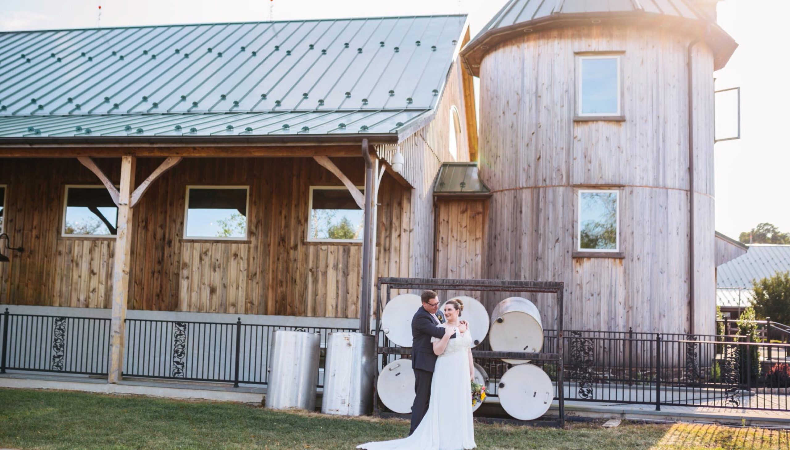 A bride and groom stand together outside a rustic wooden barn with a metal roof, next to several large barrels and metal containers.