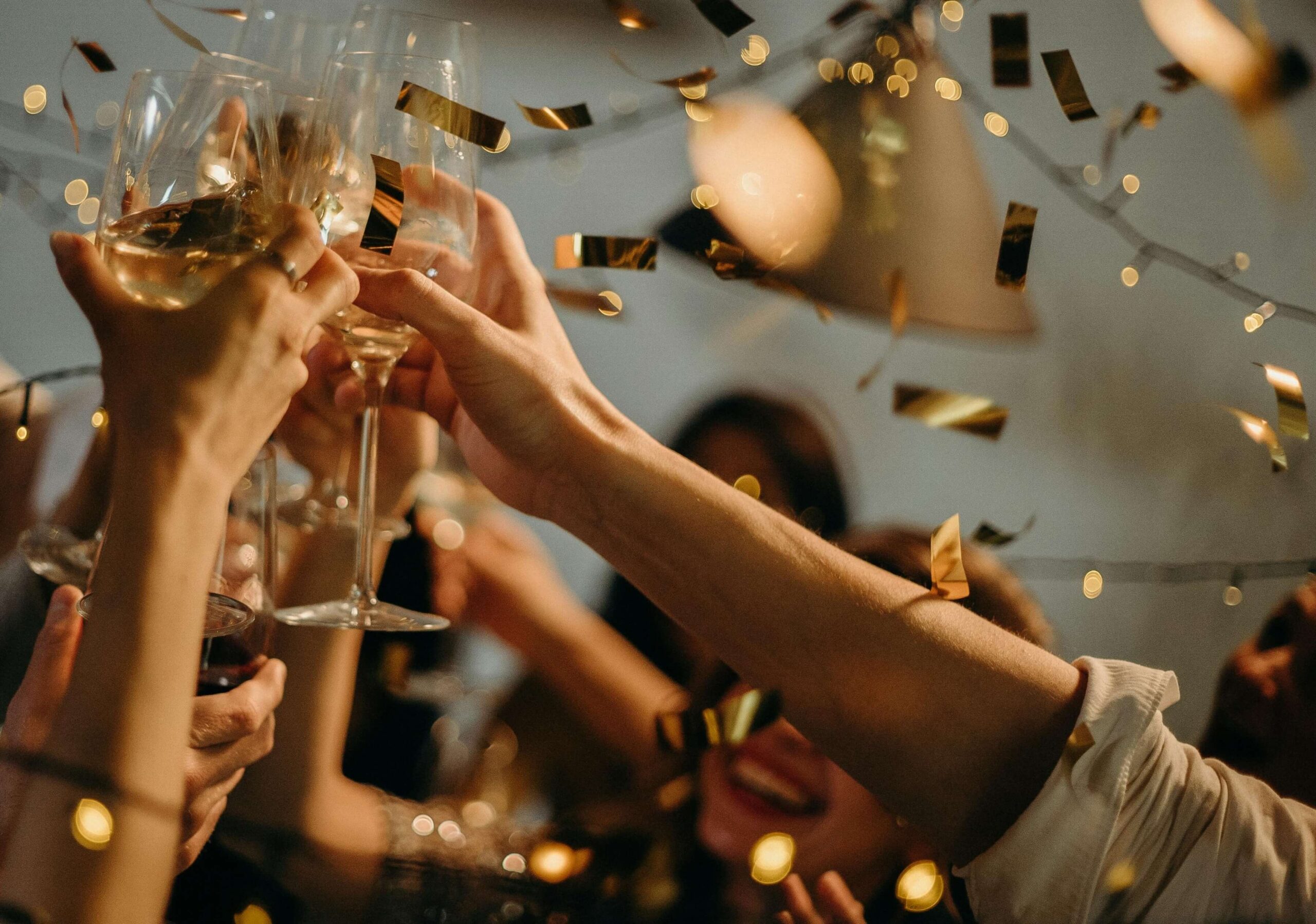 A group of people clinks glasses in a toast amid gold confetti and string lights, celebrating at an indoor gathering.